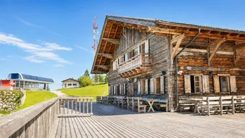 Große Holzterrasse vor einem traditionellen Holzgebäude mit Blick auf eine Seilbahnstation und Berge.