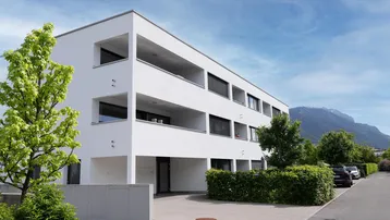 Modernes weißes Mehrfamilienhaus mit klaren Linien und Balkonen, umgeben von Grün und mit Bergblick.