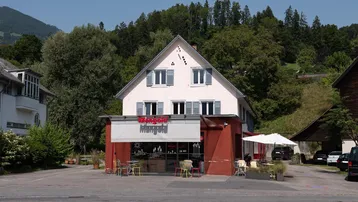 Ansicht eines Gebäudes mit Ladenfront, umgeben von Bäumen und Bergen im Hintergrund.