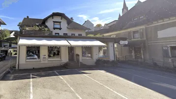Gebäude mit Geschäftsräumen im Erdgeschoss und Wohnungen darüber, umgeben von Parkplätzen und grüner Landschaft mit Kirchturm im Hintergrund.