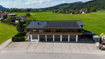 Luftaufnahme eines modernen Hauses mit Solaranlage, Garagen und weitem Blick auf grüne Felder und Berge.