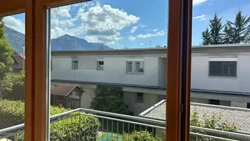 Blick vom Balkon mit Holzrahmen auf die umliegende Berglandschaft und benachbarte Gebäude.