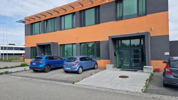 Modernes Geschäftsgebäude mit auffälliger orange-grauer Fassade und großen Fenstern, davor Parkplätze für Fahrzeuge.
