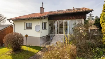 Hausfassade mit Wintergarten-Anbau, umgeben von einem kleinen Garten und Sträuchern unter blauem Himmel.