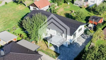Luftaufnahme eines modernen Hauses mit dunklem Dach, weißer Fassade und großem Balkon in grüner Umgebung.