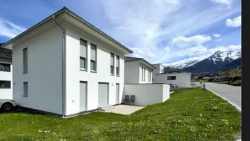 Modernes weißes Einfamilienhaus mit grüner Rasenfläche und Bergblick unter blauem Himmel.