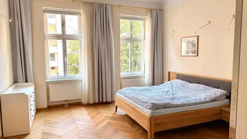 Helles Schlafzimmer mit Fischgrätparkett, Doppelbett und zwei großen Fenstern, die viel Licht hereinlassen.