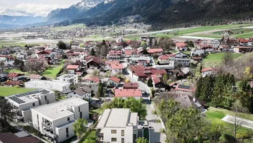 Luftaufnahme einer Wohnsiedlung mit neuen Gebäuden und umliegender Berglandschaft, umgeben von viel Grün.