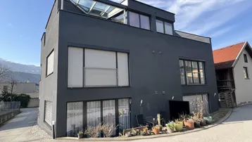 Moderne Architektur mit dunkler Fassade, großen Fenstern und Dachverglasung unter blauem Himmel.