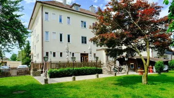 Mehrstöckiges Gebäude mit heller Fassade, umgeben von einem gepflegten Rasen und Bäumen. Eine Terrasse mit Sonnenschirmen ist sichtbar.