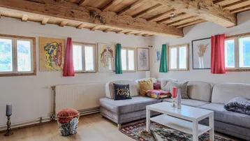 Gemütliches Wohnzimmer mit Holzbalkendecke, bequemer Couch und farbenfrohen Kunstwerken an den Wänden.