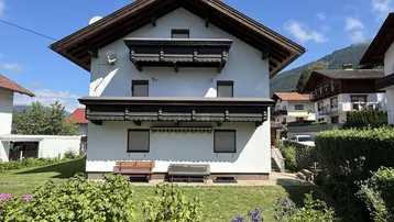 Weißes Haus mit traditionellen Holzbalkonen und gepflegtem Vorgarten unter blauem Himmel.