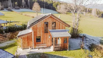 Modernes Holzhaus mit Photovoltaikanlage auf dem Dach, umgeben von grünen Wiesen und Bergen.