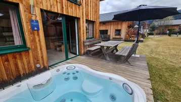 Geräumige Holzterrasse mit Whirlpool, Esstisch und Sonnenschirm vor dem Holzhaus.