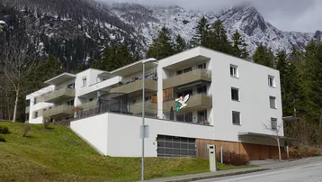 Modernes Mehrfamilienhaus mit Balkonen und Tiefgarage vor einer beeindruckenden Bergkulisse.