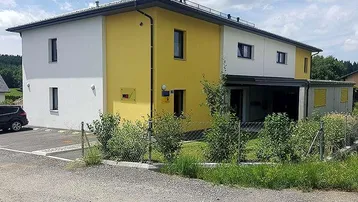 Moderne Architektur mit gelb-weißer Fassade, gepflegter Außenanlage und Parkplatzbereich.