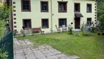 Traditionelles Haus mit hellgelber Fassade, braunen Fensterrahmen und einem gepflasterten Vorgarten mit Sitzgelegenheiten.