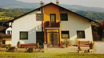Hausfassade mit gelbem Akzent und Balkon, umgeben von grüner Landschaft und Bergen.