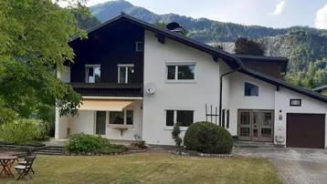 Weißes Haus mit dunklem Dach, umgeben von einem Garten und Bergen im Hintergrund.