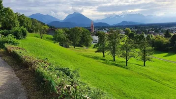 Panoramablick über grüne Hügel, ein Dorf mit Kirchturm und majestätische Berge im Hintergrund.