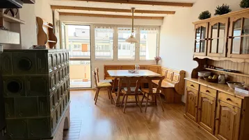 Gemütlicher Wohn-Essbereich mit traditionellem Kachelofen, Holzbalken und Zugang zum Balkon.