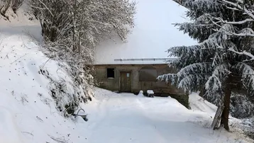 Kleines Haus im verschneiten Wald, umgeben von hohen Tannen und einer dicken Schneedecke.