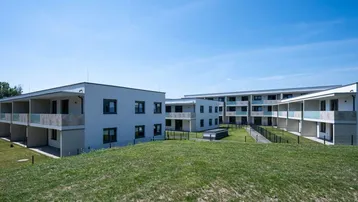 Moderne Mehrfamilienhäuser mit mehreren Wohneinheiten, Balkonen und grüner Umgebung unter blauem Himmel.