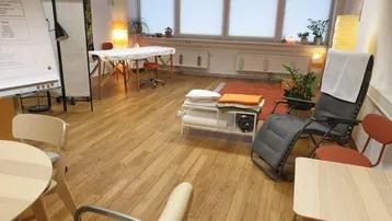 Heller Therapieraum mit Holzfußboden, großen Fenstern und verschiedenen Sitzgelegenheiten für Behandlungen und Beratungen.