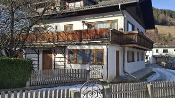 Traditionelles Wohnhaus mit Holzbalkonen und Satteldach, umgeben von winterlicher Landschaft und Bergen.