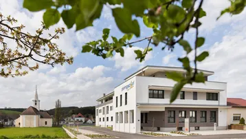 Modernes Gebäude mit klarer Architektur und Blick auf eine Kirche im Grünen.