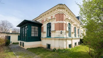 Historisches Gebäude mit markanter Architektur, einer verglasten Veranda und gepflastertem Weg im Vorgarten.