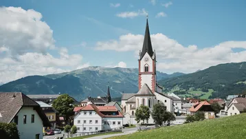 Malerisches Dorf mit Kirche und Bergen im Hintergrund unter blauem Himmel.