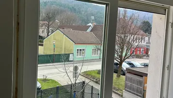 Blick aus einem Fenster auf eine ruhige Wohnstraße mit Häusern, Bäumen und entfernten Hügeln.