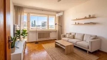 Helles Wohnzimmer mit beigem Sofa, Holzboden und großem Fenster mit Blick ins Grüne.