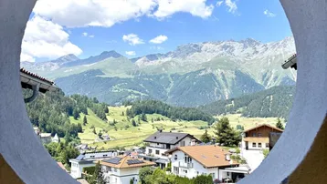 Malerischer Bergblick durch ein rundes Fenster, das eine idyllische Alpenlandschaft mit Häusern zeigt.