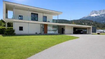 Moderne Villa mit Flachdach, großen Glasflächen, Carport und Bergblick unter strahlend blauem Himmel.