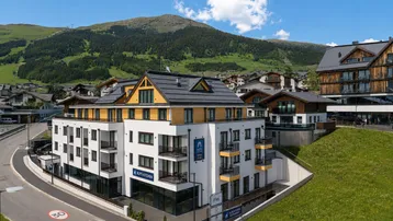 Modernes Gebäude in alpiner Umgebung mit Balkonen und Bergblick unter blauem Himmel.