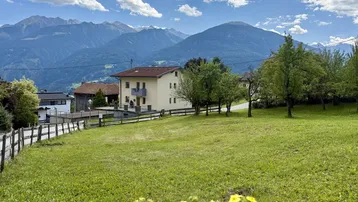Haus in ländlicher Umgebung mit Bergen im Hintergrund und grüner Wiese im Vordergrund.