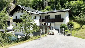 Traditionelles Haus in den Bergen mit weißer Fassade und dunklem Dach, umgeben von viel Grün.