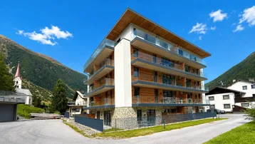 Modernes Apartmentgebäude mit Holzfassade und Glasbalkonen, umgeben von alpiner Berglandschaft.