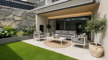 Moderne Terrasse mit bequemen Gartenmöbeln, Rasenfläche und Steinmauer, ideal zum Entspannen im Freien.