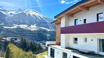 Modernes Gebäude mit Balkon und Terrasse, eingebettet in eine beeindruckende Berglandschaft unter blauem Himmel.