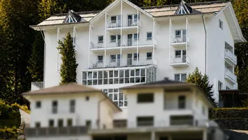 Weißes Mehrfamilienhaus in den Bergen mit vielen Balkonen und umgeben von dichtem Wald.