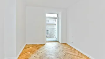 Helles Zimmer mit Fischgrätparkett und Zugang zu einem französischen Balkon, bietet viel Tageslicht.