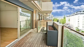 Geräumiger Balkon mit Holzboden, Glasgeländer und Blick auf die umliegenden Gebäude und den Himmel.