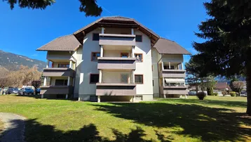 Mehrfamilienhaus mit mehreren Balkonen und gepflegter Grünfläche im Vordergrund unter blauem Himmel.