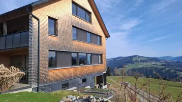 Modernes Holzhaus mit Schindelfassade und weitem Blick auf die Berge und grüne Hügel.