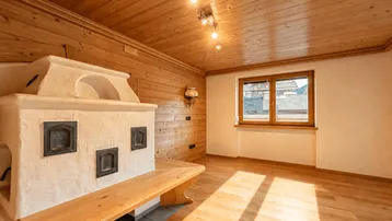 Helles Zimmer mit Holzboden, Holzdecke und einem traditionellen Kachelofen, der Wärme spendet.