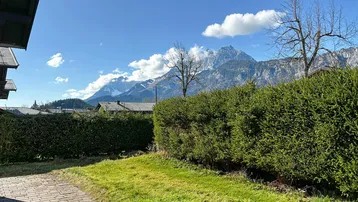 Grüner Gartenbereich mit gepflastertem Weg, Hecke und beeindruckendem Bergblick unter blauem Himmel.