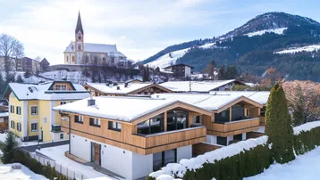 Moderne Gebäude in einer verschneiten Alpenlandschaft mit traditioneller Kirche im Hintergrund.
